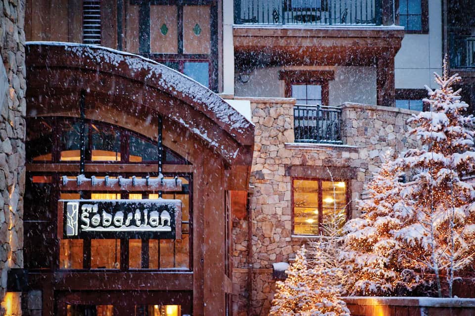Snowy alpine village street scene with Sebastien restaurant storefront lit up in evening, evergreen trees, and snow-covered buildings