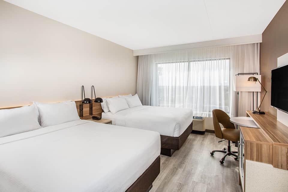Contemporary hotel room with two queen beds, TV, and sheer curtains by window