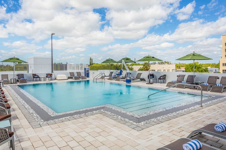 Outdoor swimming pool with lounge chairs, green umbrellas and landscaped resort grounds under blue sky