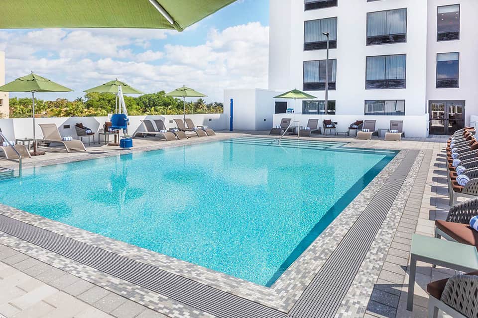 Resort pool deck with turquoise water, green umbrellas, lounge chairs, and white hotel building