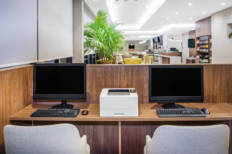 Modern hotel business center with dual monitors, keyboard, printer, and potted plant decor