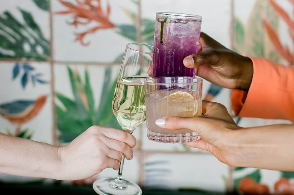 Three people clinking glasses with wine, water, and purple cocktail against a tropical botanical wallpaper backdrop
