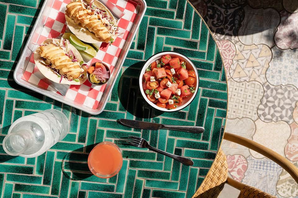 Overhead view of meal on green herringbone table: lobster rolls, salad bowl, water glass, and coral drink.