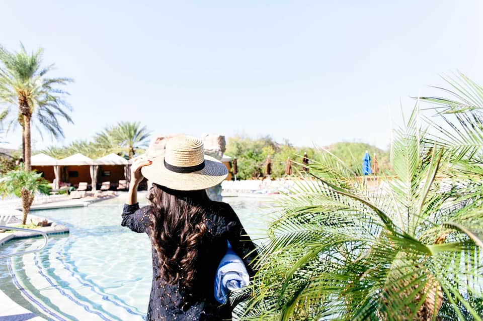 Person in straw hat viewing resort pool with palm trees and thatched-roof cabanas