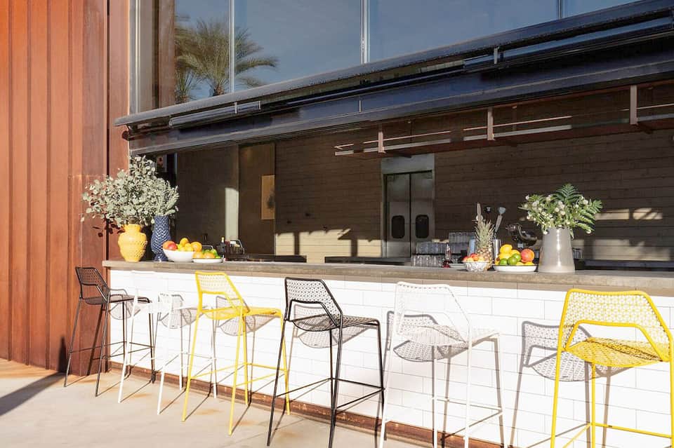 Outdoor bar counter with yellow and neutral modern bar stools, palms, and large windows