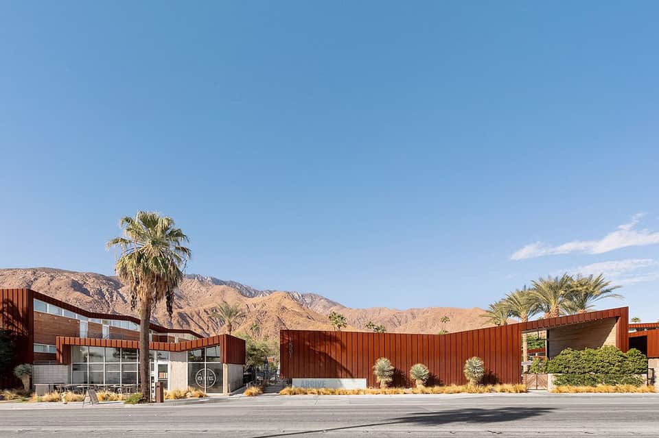Modern resort building with corrugated metal facade and palm trees against desert mountains
