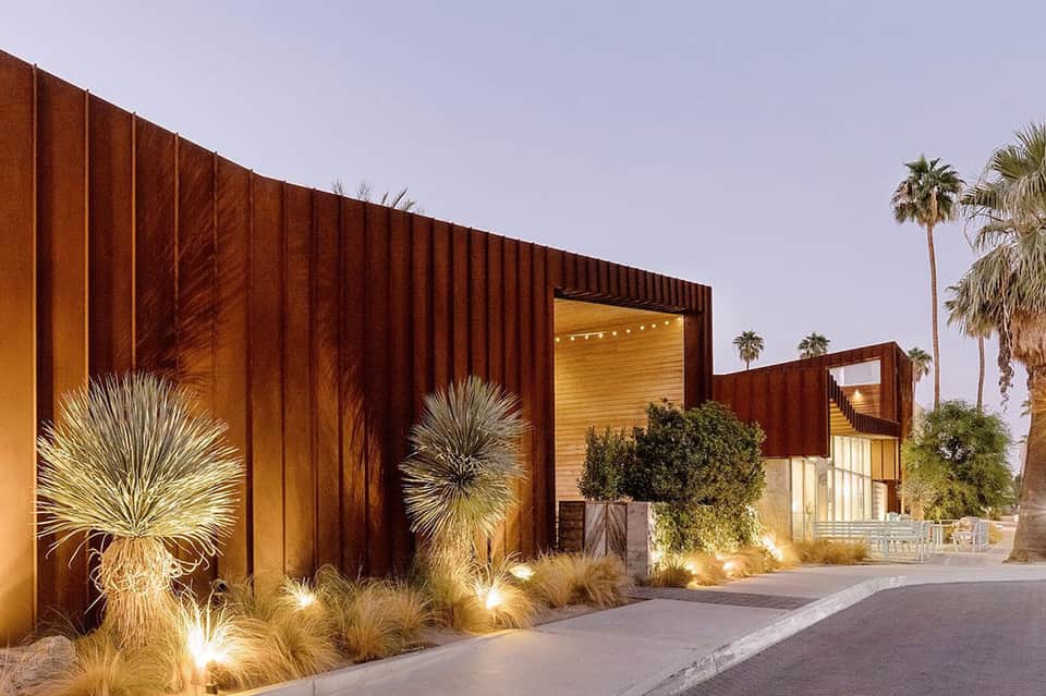 Contemporary entrance with corrugated metal walls and palm trees illuminated at dusk