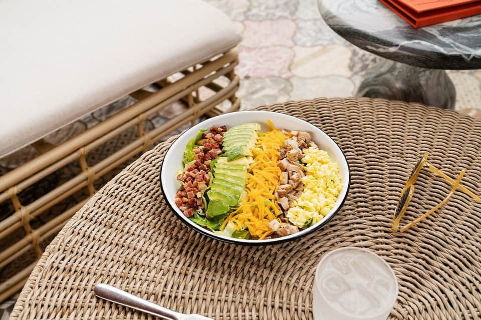 Salad bowl with avocado, bacon, egg, and cheese on woven table with resort lounge chairs in background
