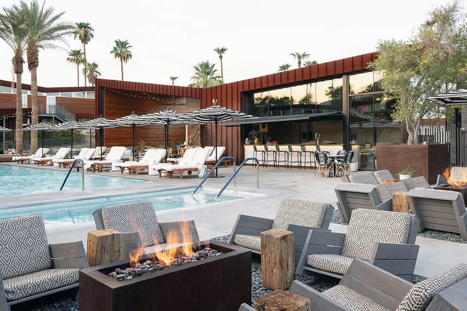 Modern resort pool area with fire pit, lounge seating, umbrellas, glass building, and palm tree landscaping