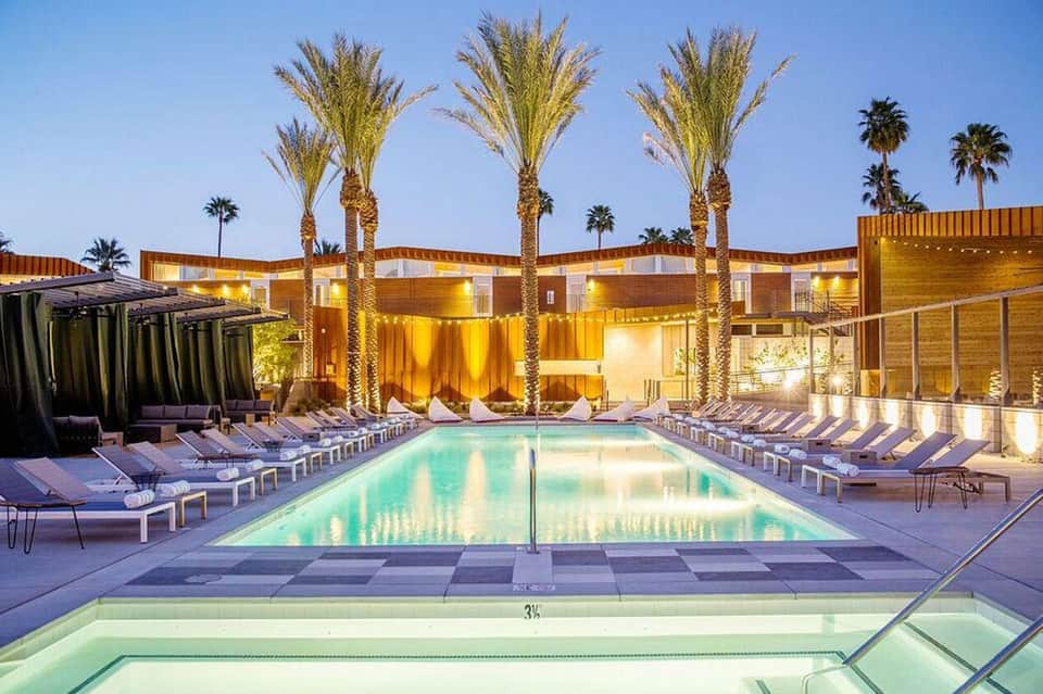 Resort pool at dusk with glowing buildings, palm trees, lounge chairs, and illuminated landscaping