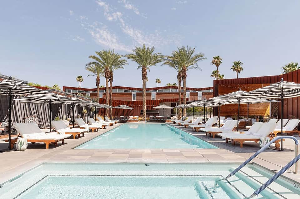 Resort pool with palm trees, white loungers, striped umbrellas, and rust-colored building