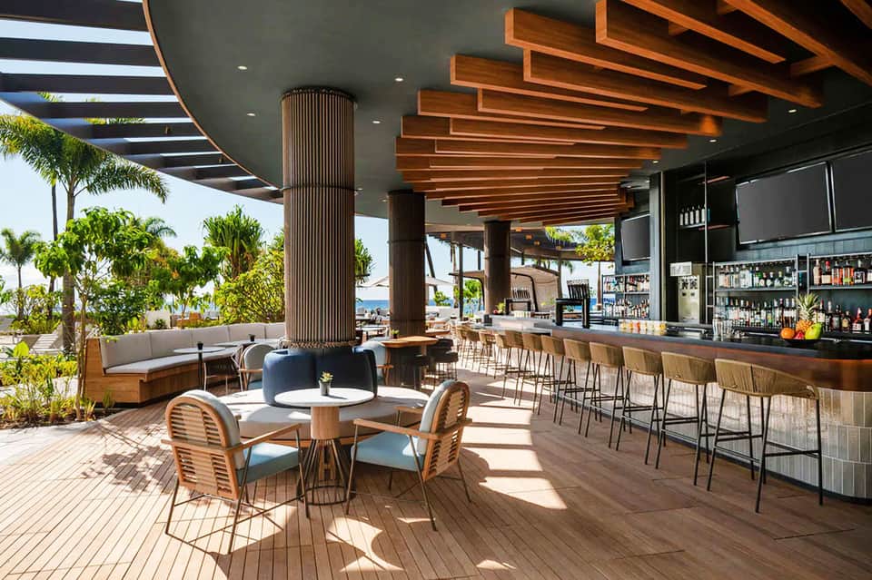 Elevated rooftop bar with wood decking, wooden furniture, tropical plants, and water views with palm trees
