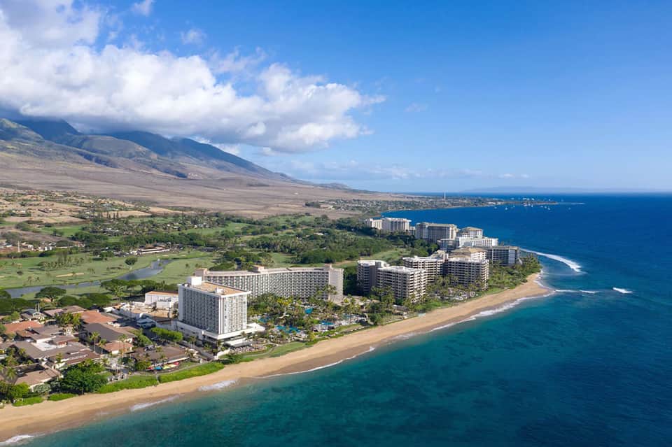 Aerial view of beachfront resort with multiple high-rise hotels, sandy beach, and mountain backdrop in Hawaii