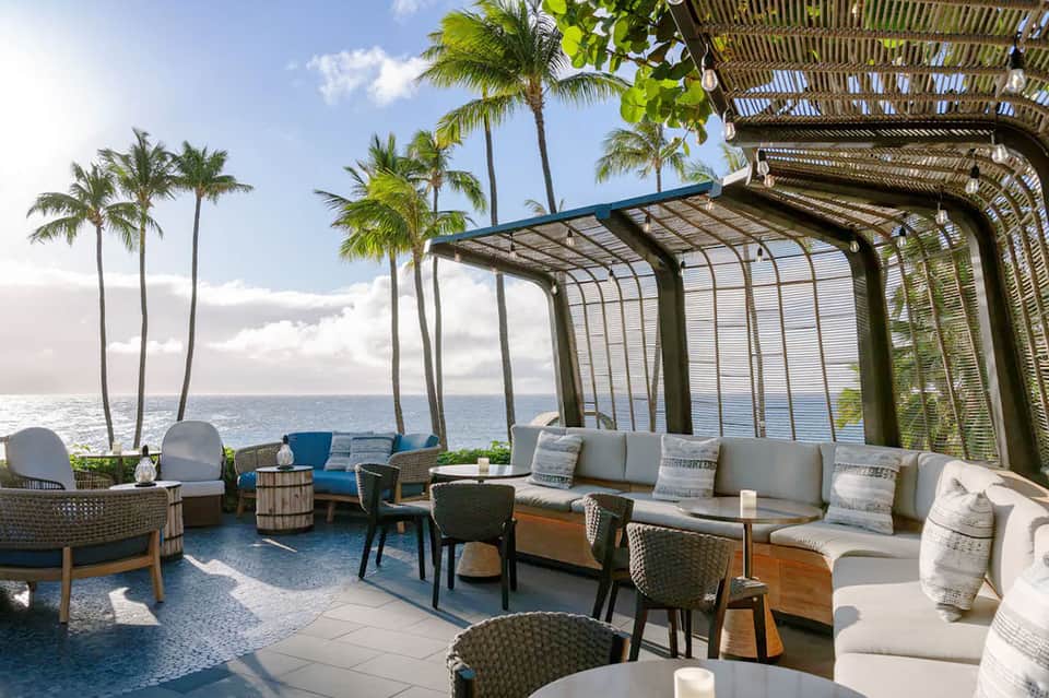 Beachfront lounge area with pergola, palm trees, ocean view, and gray cushioned seating