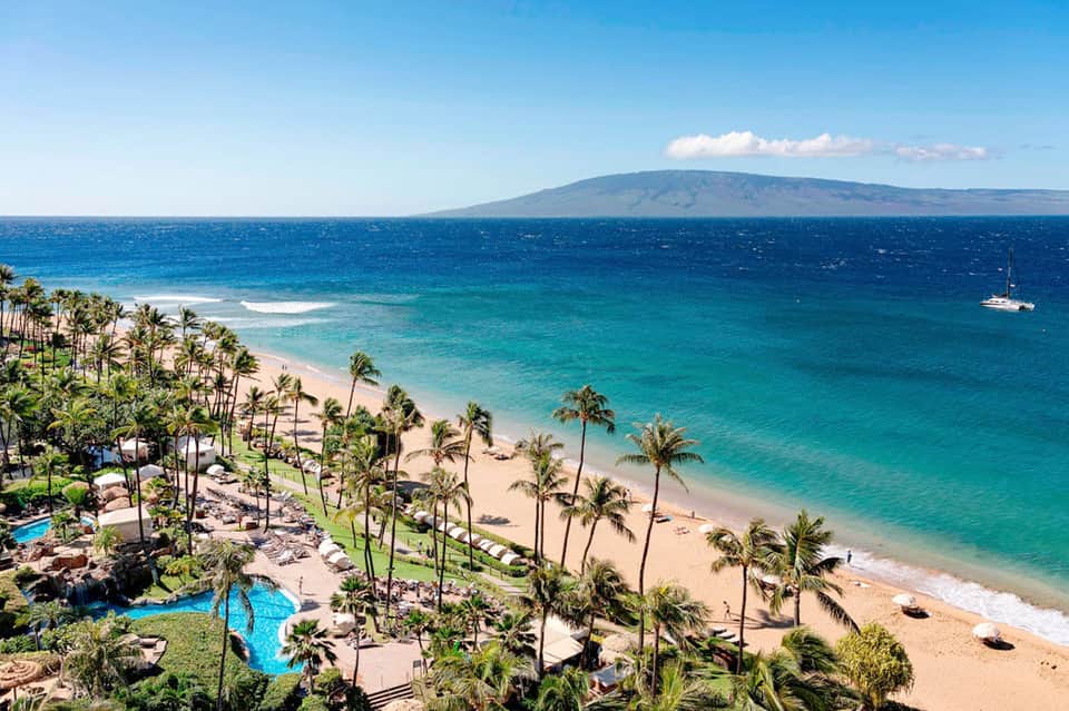 Aerial beach resort view with palm trees, turquoise ocean, and distant volcanic mountain