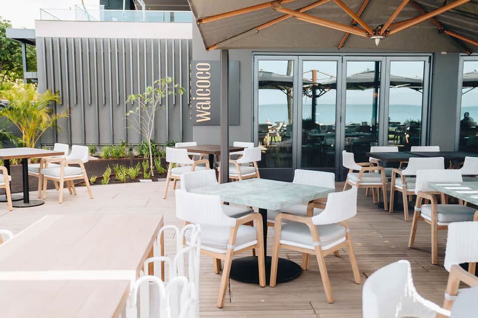 Modern beachfront restaurant terrace with wooden furniture, white umbrellas, ocean views, and contemporary design