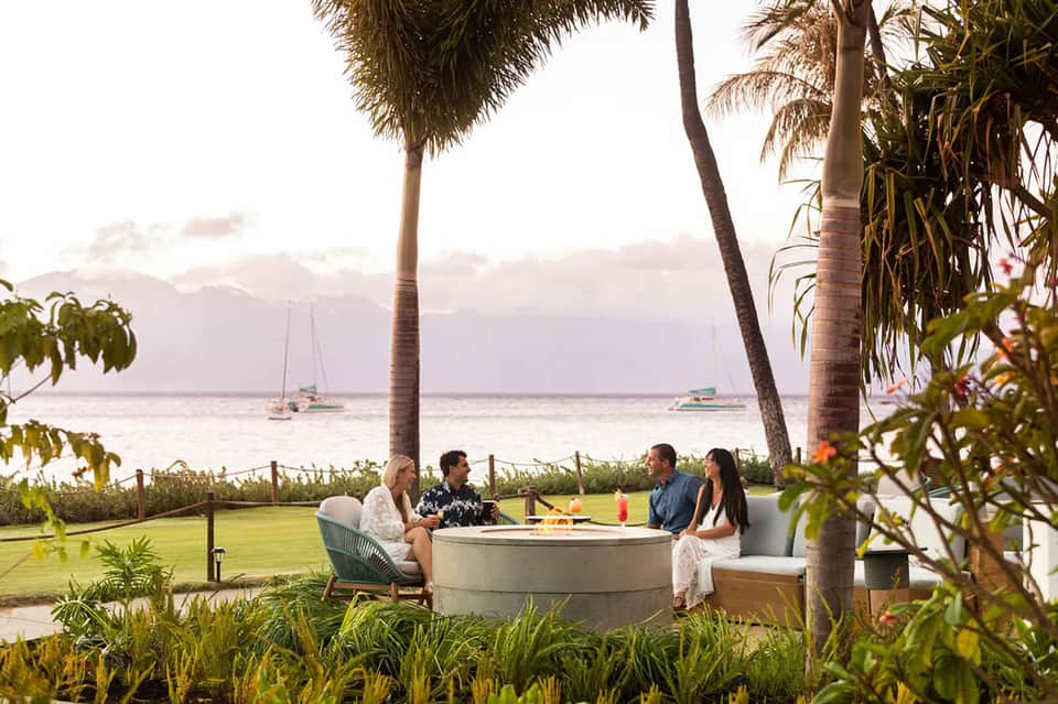 Four guests relaxing around a fire pit table on a manicured lawn overlooking sailboats and ocean