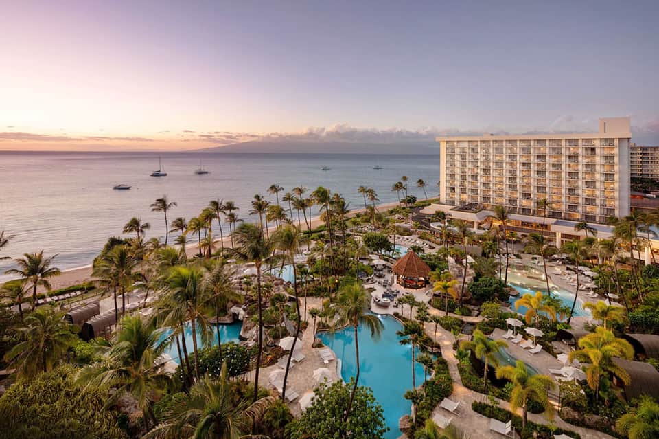Aerial view of oceanfront resort at sunset with pools, palm trees, beach, and sailboats