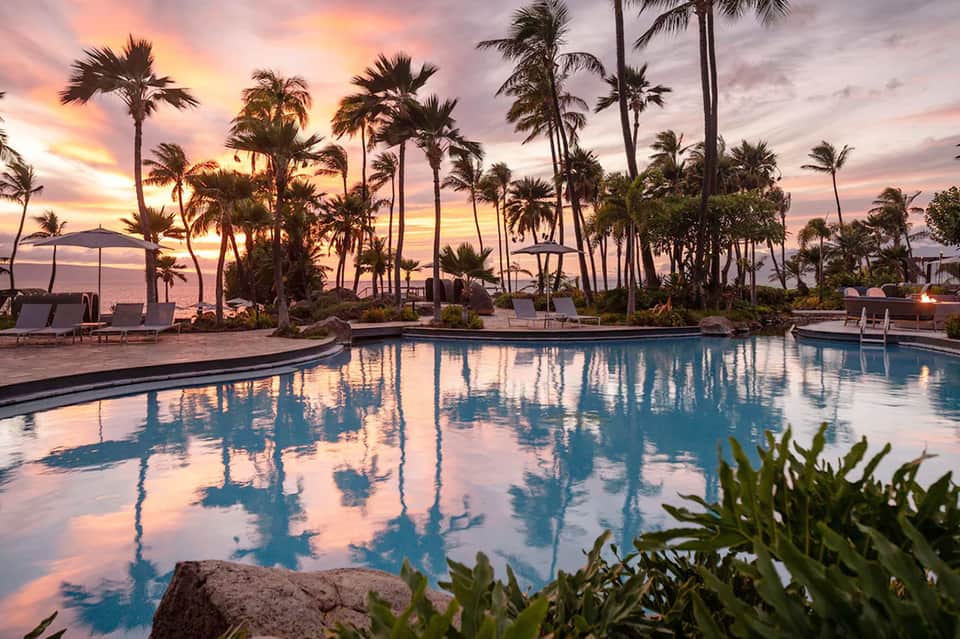 Resort pool at sunset with palm trees silhouetted against pink and purple sky, lounge chairs nearby