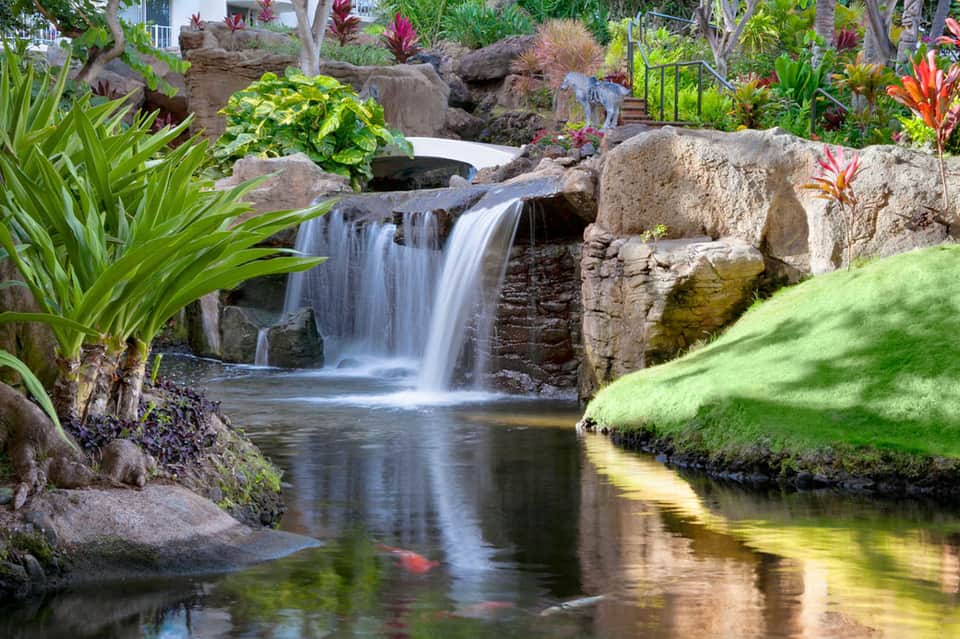 Landscaped garden waterfall cascading over rocks with tropical plants, flowers, and manicured grass