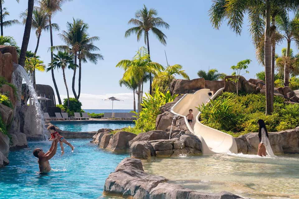 Lagoon-style resort pool with water slides, tropical landscaping, and palm trees beside the ocean