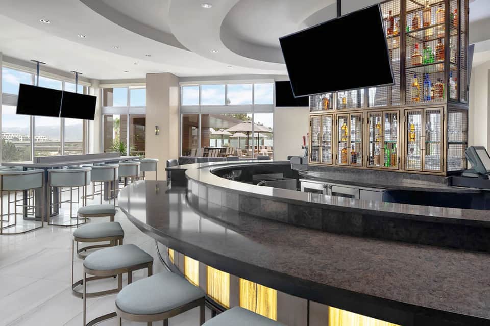 Modern hotel lobby bar with curved black counter, flat-screen TVs, and beverage display shelving