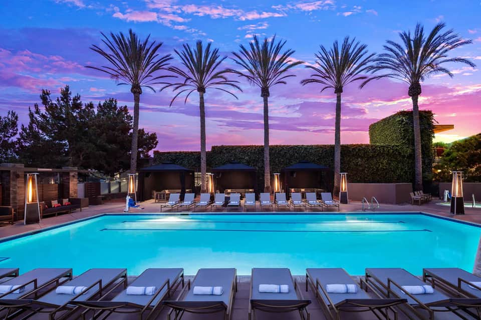 Resort pool at dusk with palm trees, lounge chairs, and glowing heaters