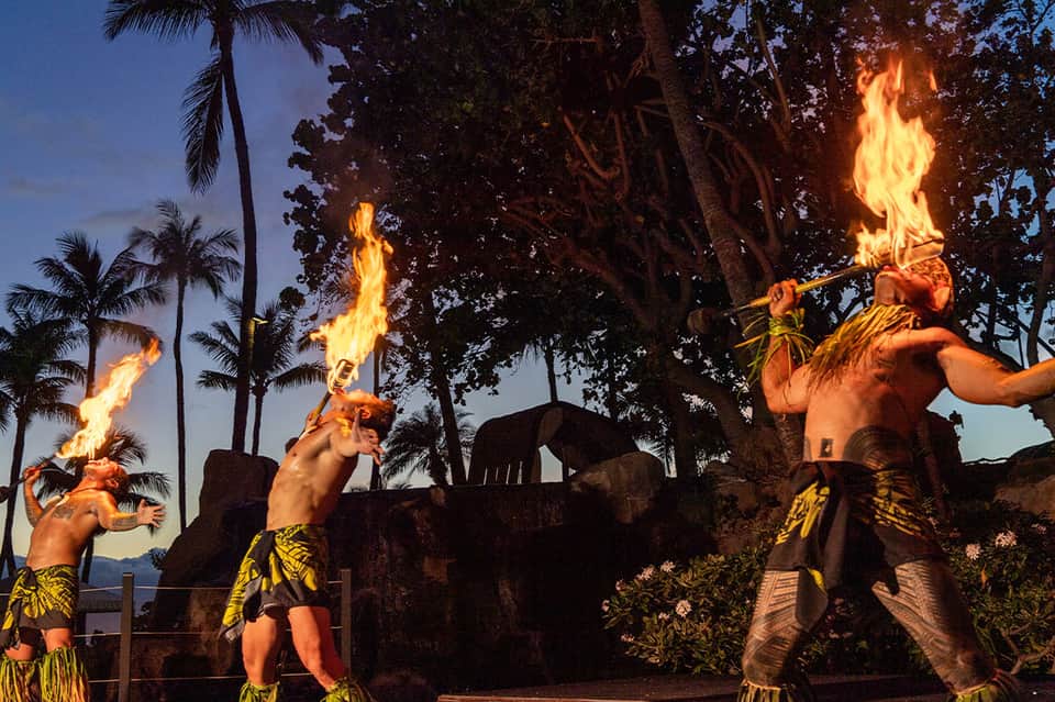 Fire performers in traditional Hawaiian attire performing fire torch dance at dusk with palm trees and ocean view