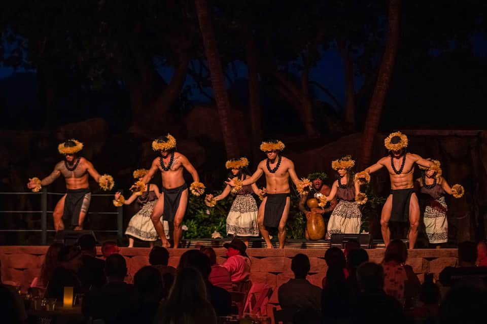 Traditional Hawaiian luau performance with shirtless male dancers in straw skirts on lit stage