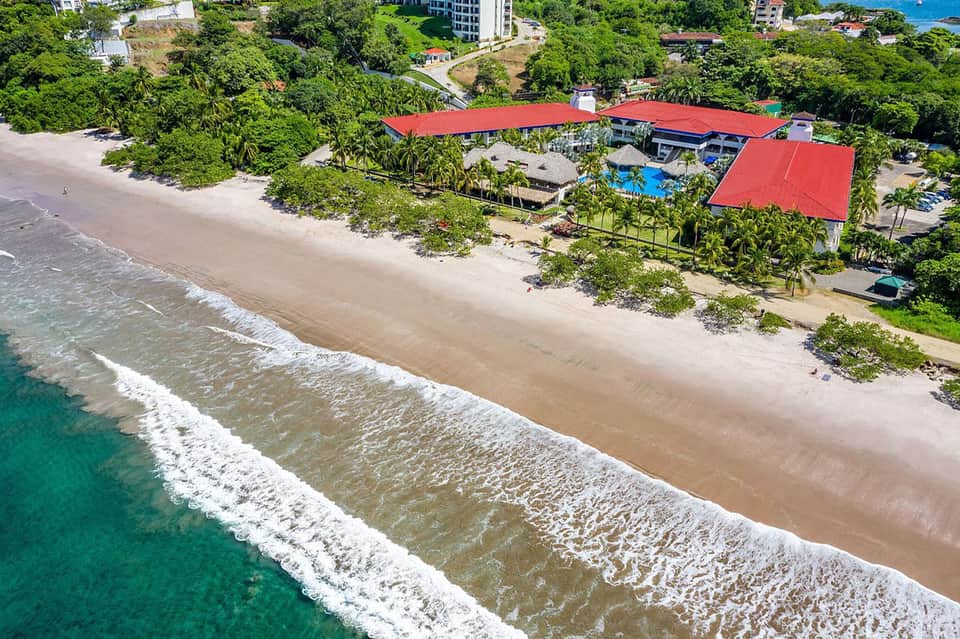 Aerial view of beachfront resort with red-roofed structures, blue pool, palm trees, and pristine sandy beach