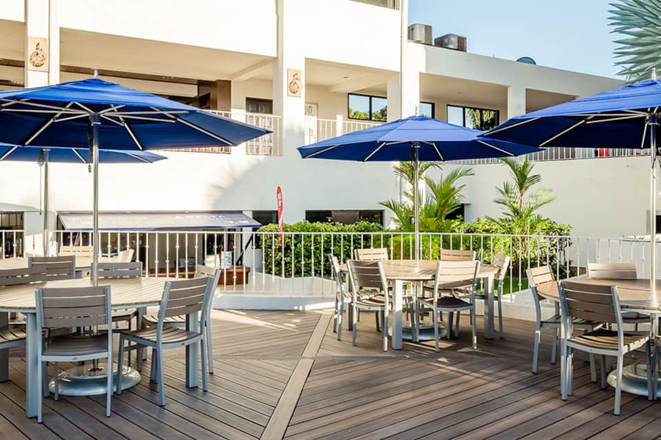 Outdoor deck with dining tables, blue umbrellas, lounge chairs, and modern white building