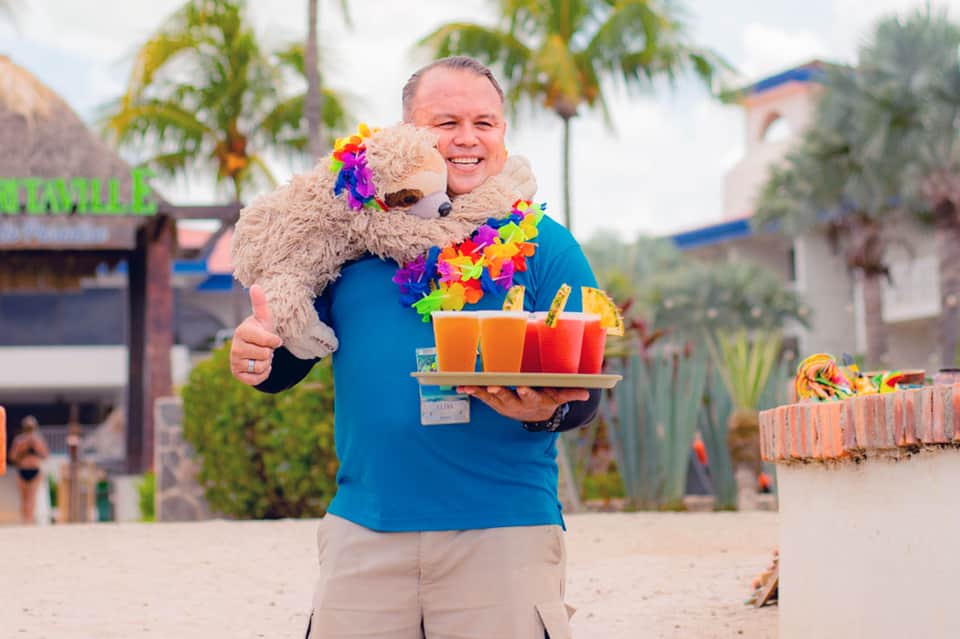 Staff member holding alpaca and tropical drinks on beach with palm trees