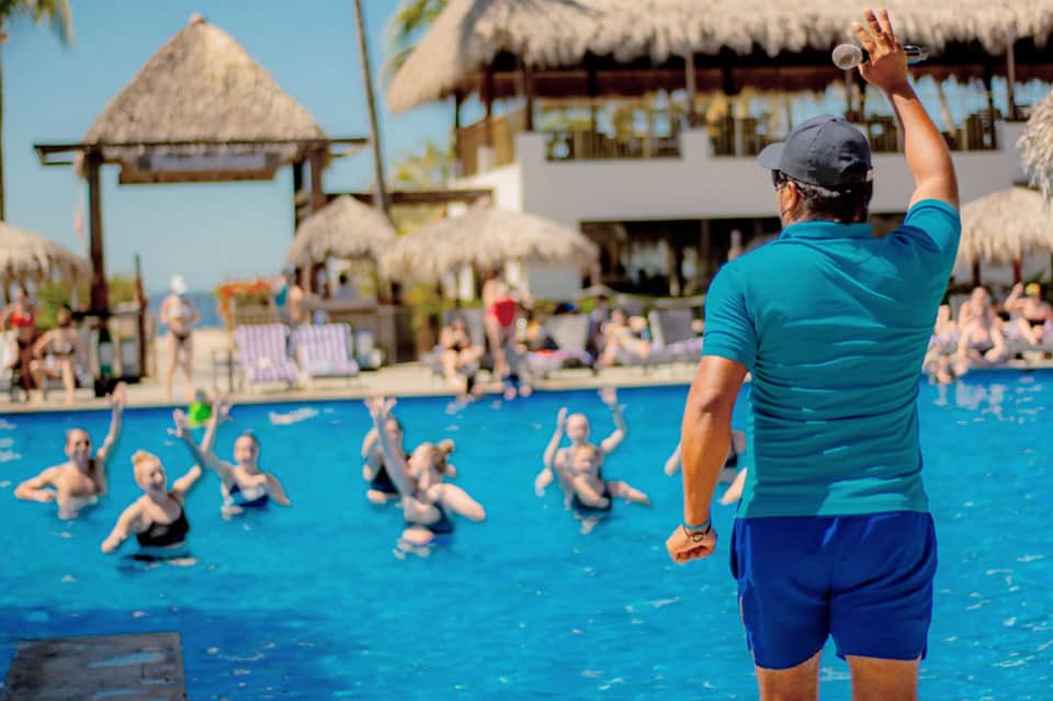 Fitness instructor leading water aerobics class in bright blue pool with tropical thatch structures
