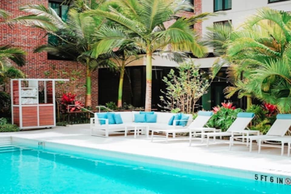 Resort pool area with white lounge chairs, tropical palm trees, and brick hotel building