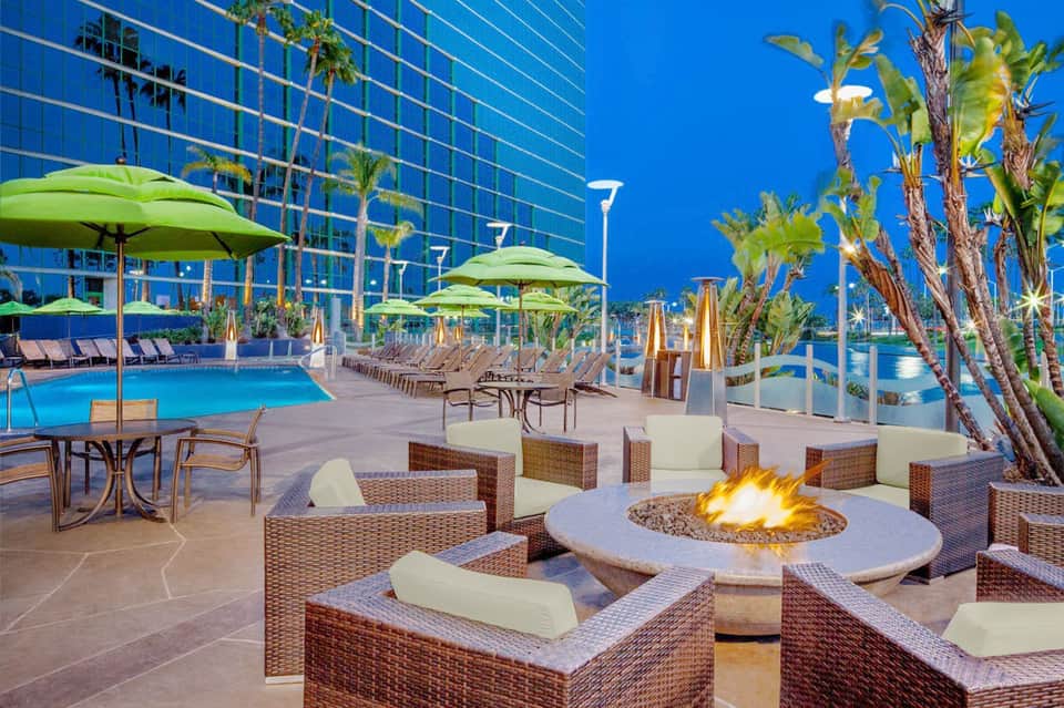 Luxury resort pool deck with fire pit, wicker lounge seating, palm trees, and modern high-rise hotel building
