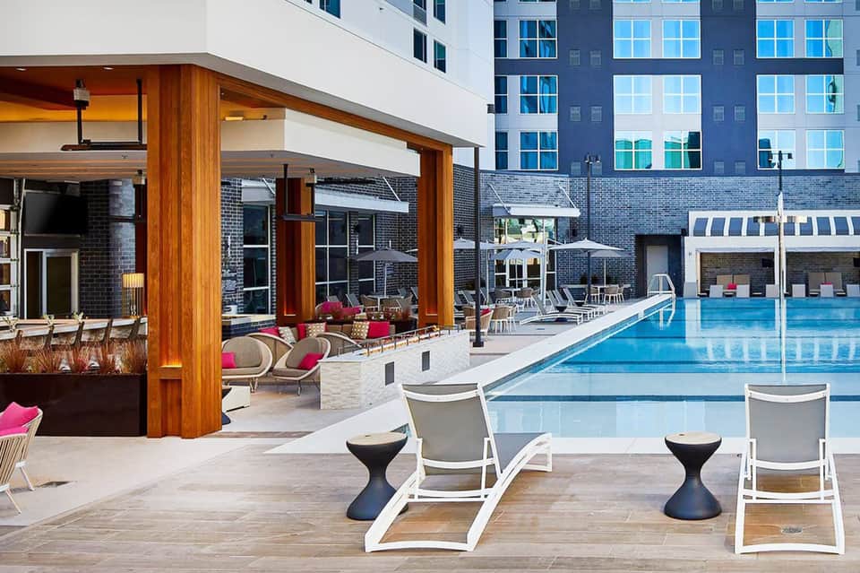 Resort pool area with lounge chairs, modern wooden pergola structure, pink accent furnishings, and multi-story hotel building backdrop