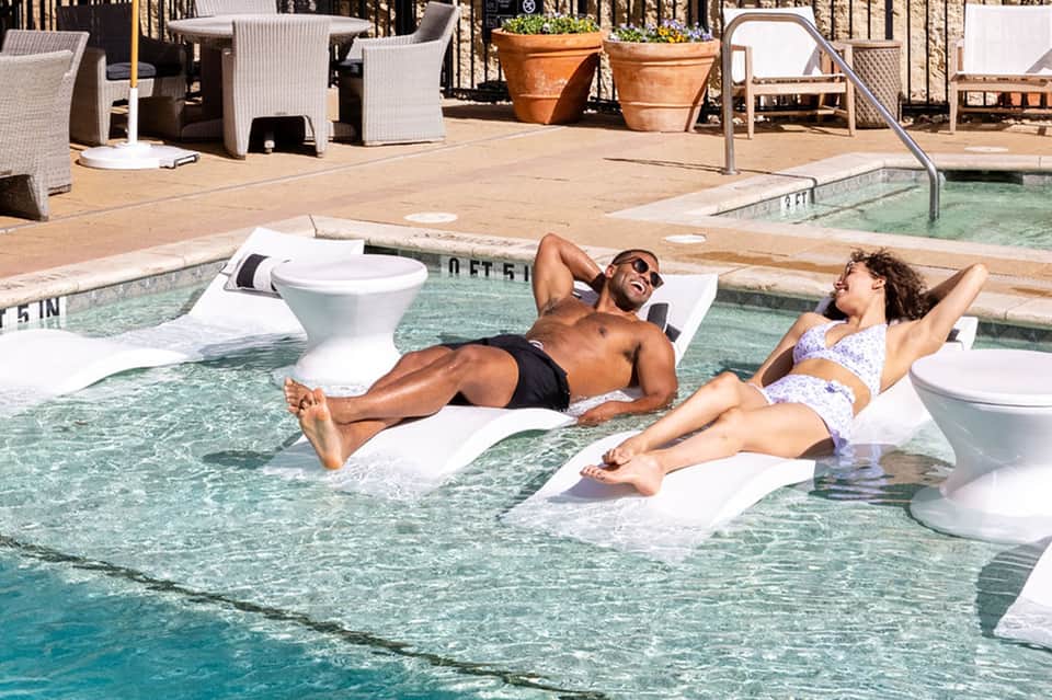 Two guests floating on white loungers in shallow pool with white furniture and railings