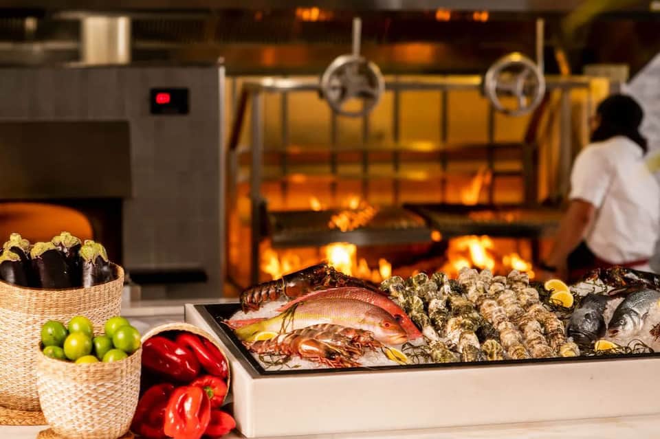 Open kitchen with fireplace, fresh seafood display, vegetables, and chef preparing food