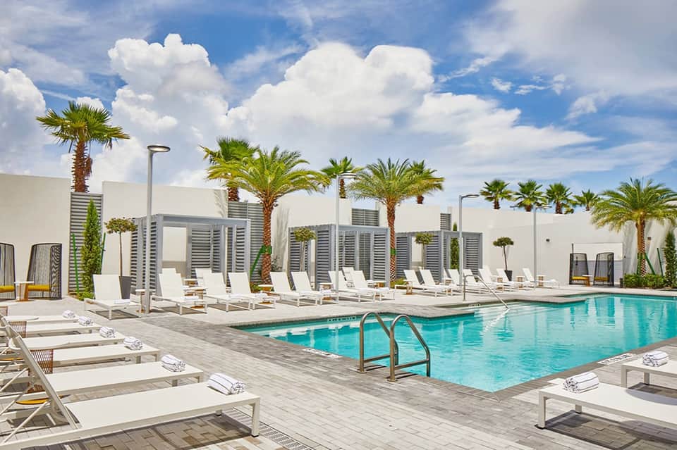 Modern resort pool deck with white lounge chairs, palm trees, white architecture, and clear blue sky with clouds.