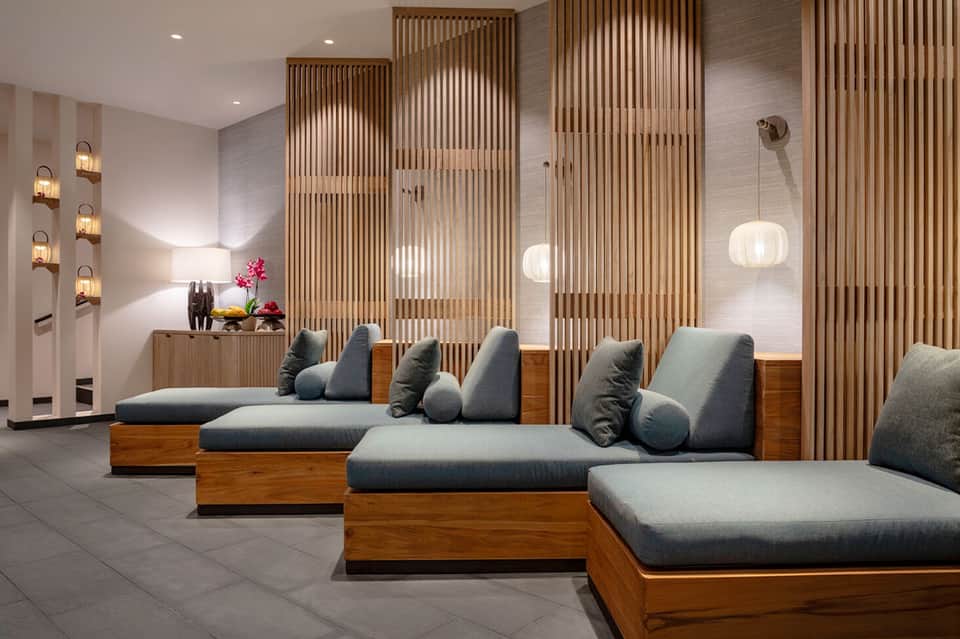 Spa relaxation area with wooden loungers, gray cushions, decorative wall sconces, and wood slats