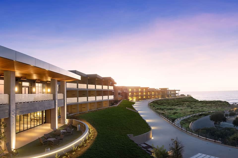 Modern curved resort buildings on grassy hillside at twilight with ocean view and lit pathways