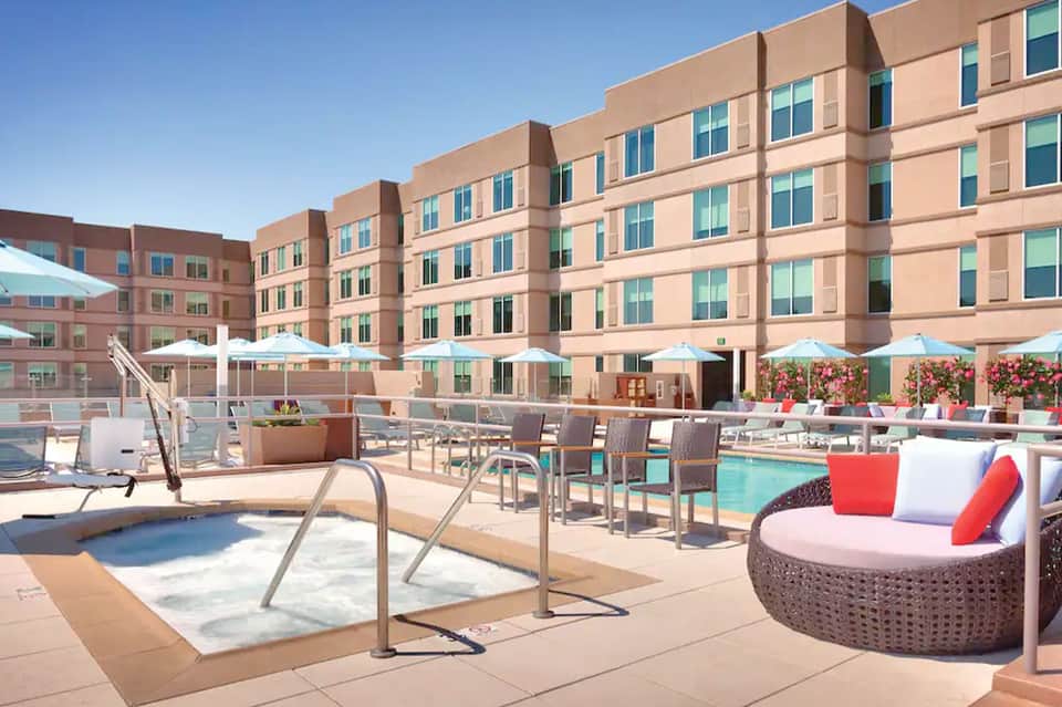 Resort courtyard pool area with lounge chairs, umbrellas, modern pink building, and sunny day