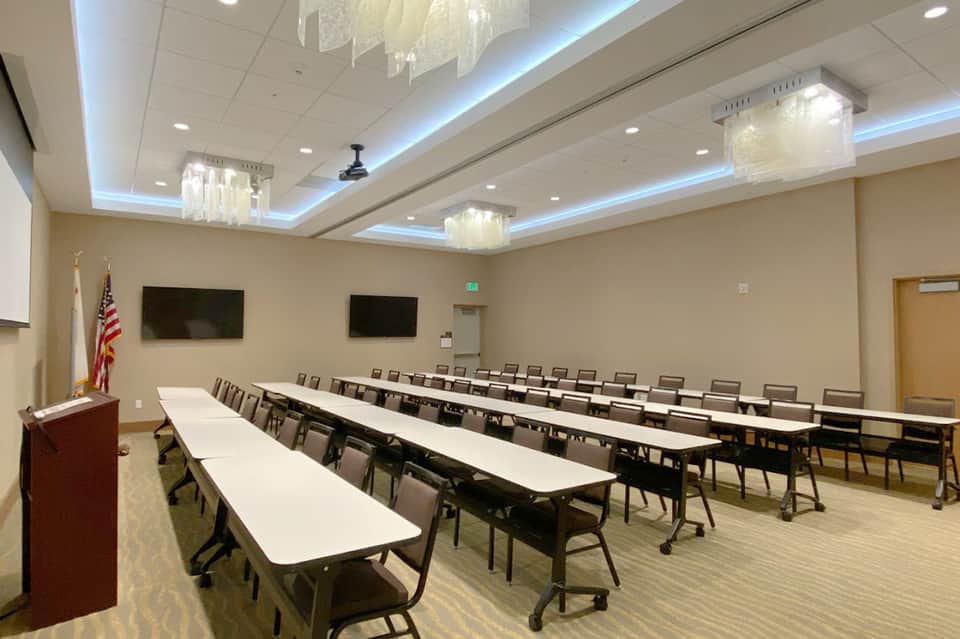 Spacious ballroom with long conference tables, chairs, ceiling fixtures, and flat-screen displays