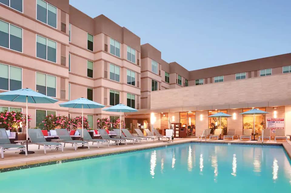 Resort pool at dusk with pink building, lounge chairs, and blue umbrellas