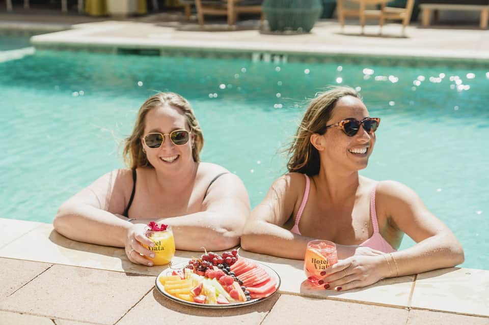 Two women in pool holding tropical drinks and fresh fruit platter at poolside