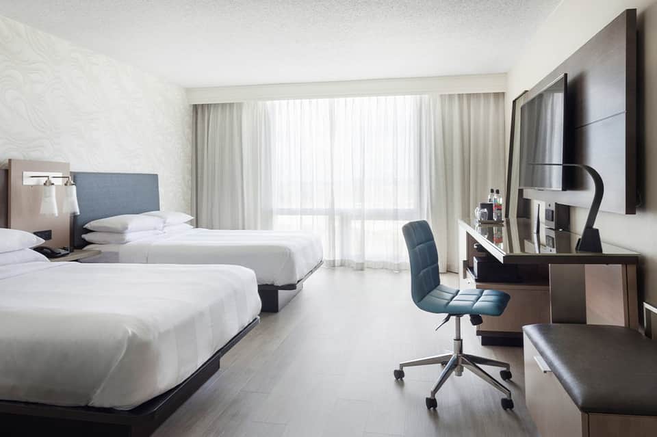 Modern hotel room with two queen beds, desk workspace, flat-screen TV, and balcony access