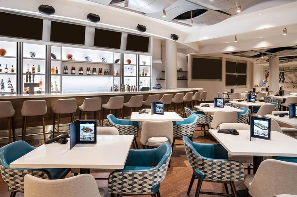 Modern hotel dining restaurant with illuminated bar shelving, geometric patterned chairs, and tablets