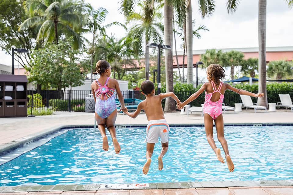 Three children jumping into resort pool with palm trees and lounge chairs visible poolside