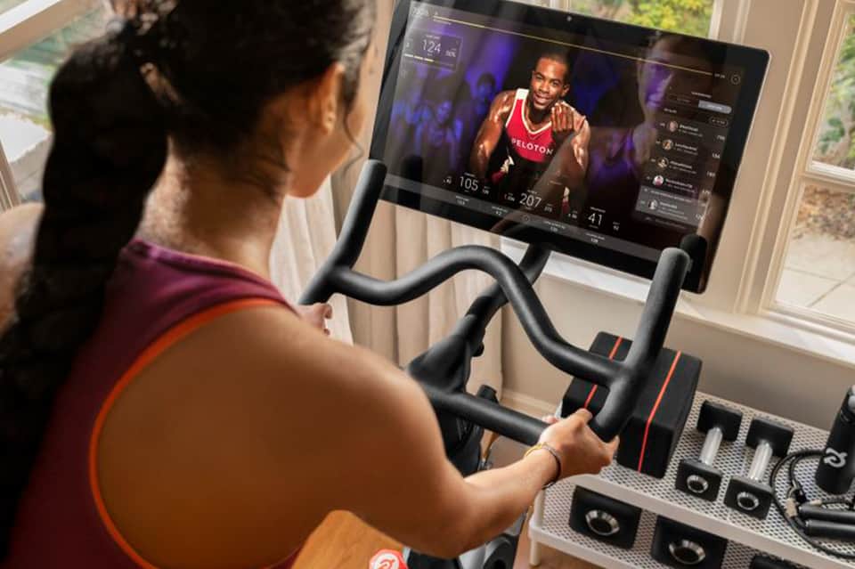 Woman on stationary bike following live fitness instructor on tablet screen