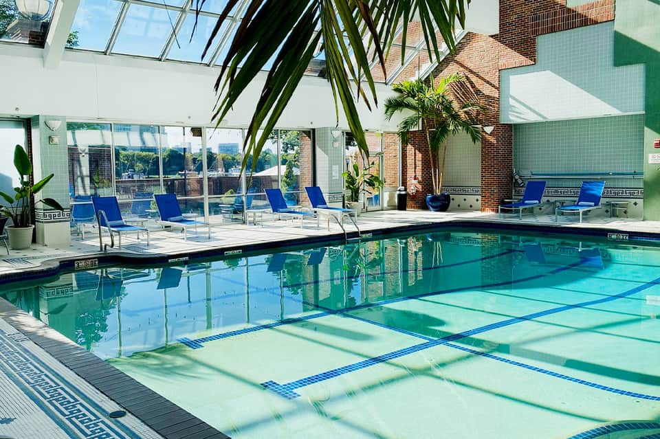 Indoor lap pool with lane dividers, large windows, blue lounge chairs, and tropical plants